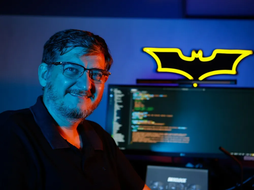Aislan Greca sorrindo, com um letreiro de neon do Batman e um monitor de código ao fundo